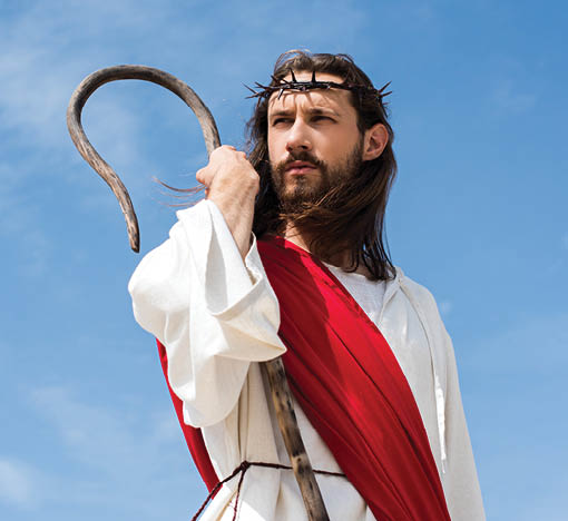 Jesus in robe, red sash and crown of thorns standing with wooden staff against blue sky and looking away