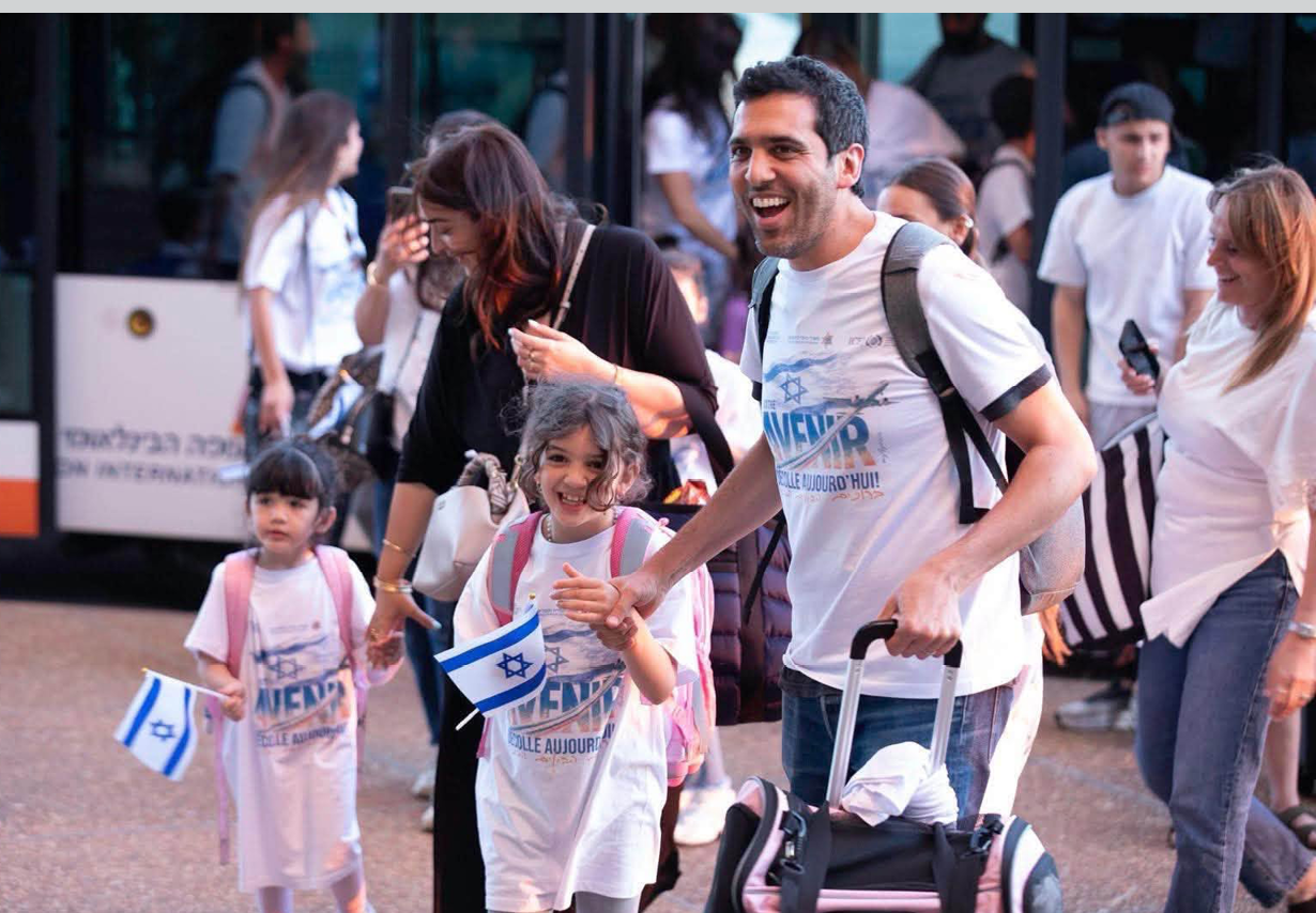 A smiling child with curly hair and a backpack is holding hands with an adult wearing a white shirt with blue text and symbols. Description generated by AI